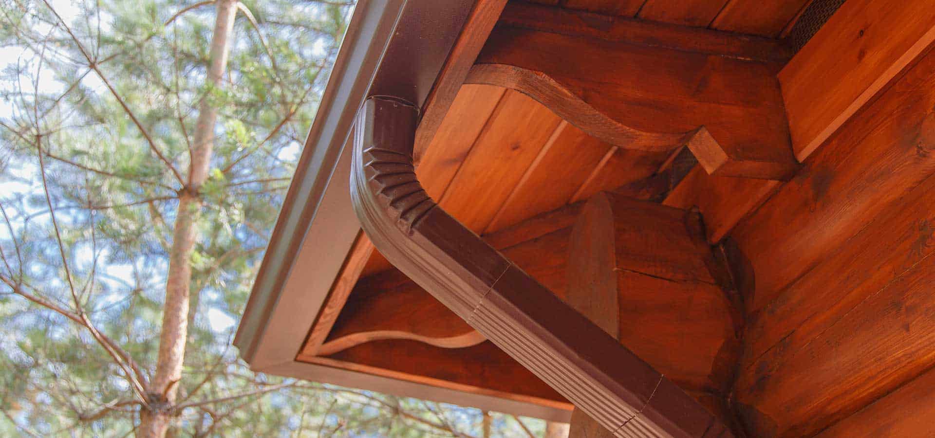 Close-up view of a brown rain gutter and downspout attached to the roof of a wooden structure, with trees and sky visible in the background.